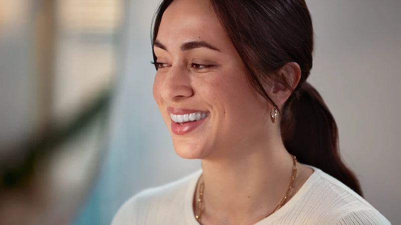 Woman with a smile, wearing a white top and gold necklace, against a blurred indoor background.